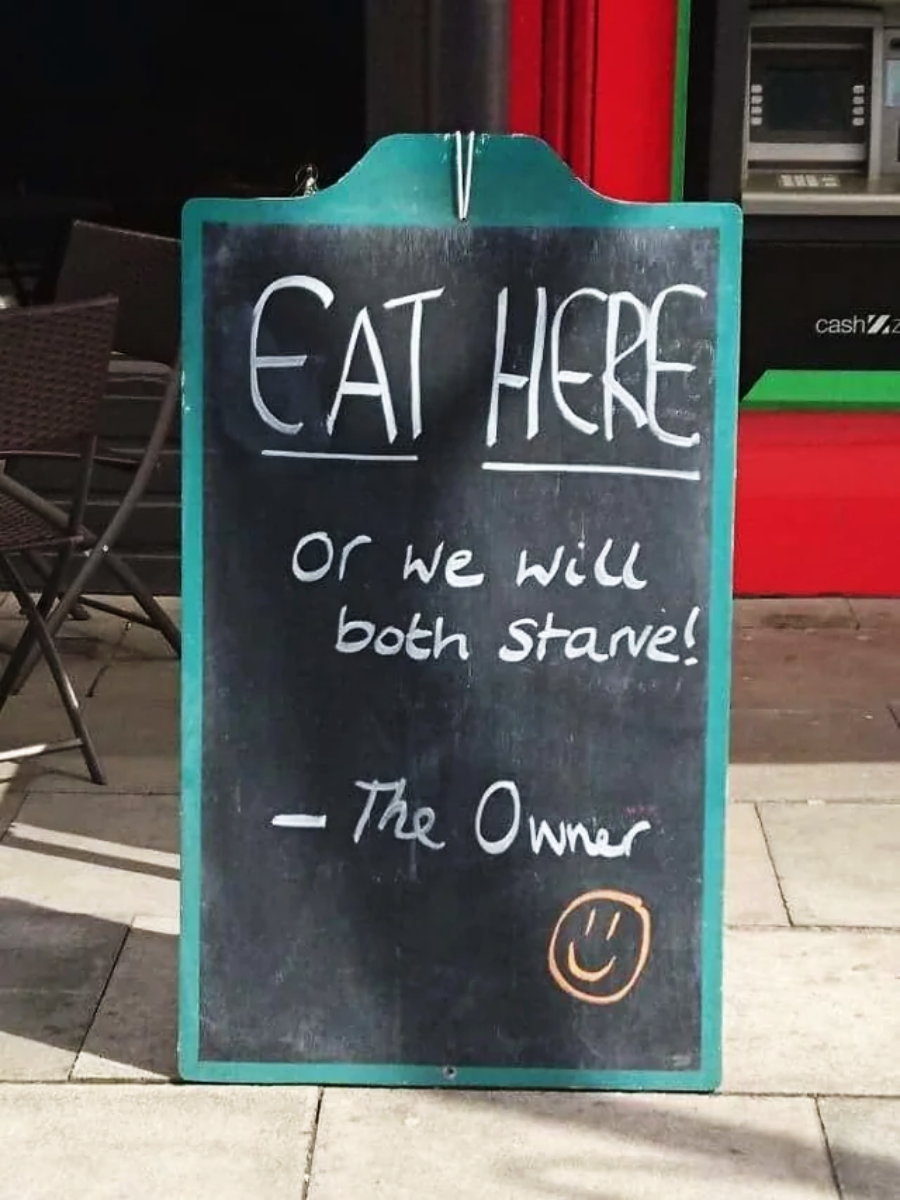 A chalkboard sign on a sidewalk reads: “EAT HERE or we will both starve! – The Owner” with a drawn smiley face at the bottom. An ATM and cafe chairs are visible in the background.