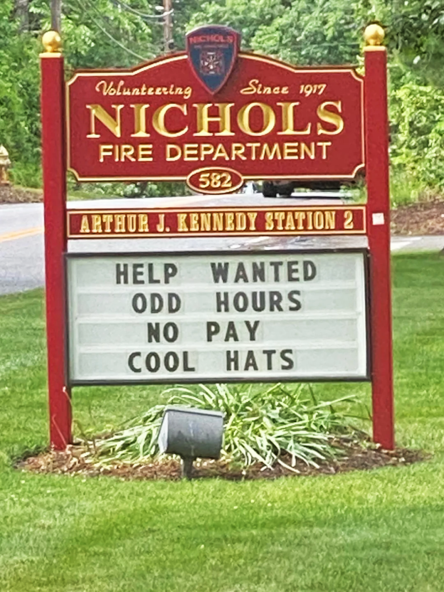 A red Nichols Fire Department sign reads: "HELP WANTED ODD HOURS NO PAY COOL HATS," with greenery and a road in the background.