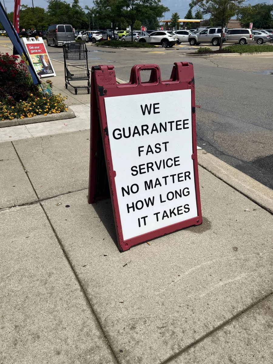 A sidewalk sign with bold black text reads: "WE GUARANTEE FAST SERVICE NO MATTER HOW LONG IT TAKES." The sign is on a concrete walkway near a parking lot and flowers.