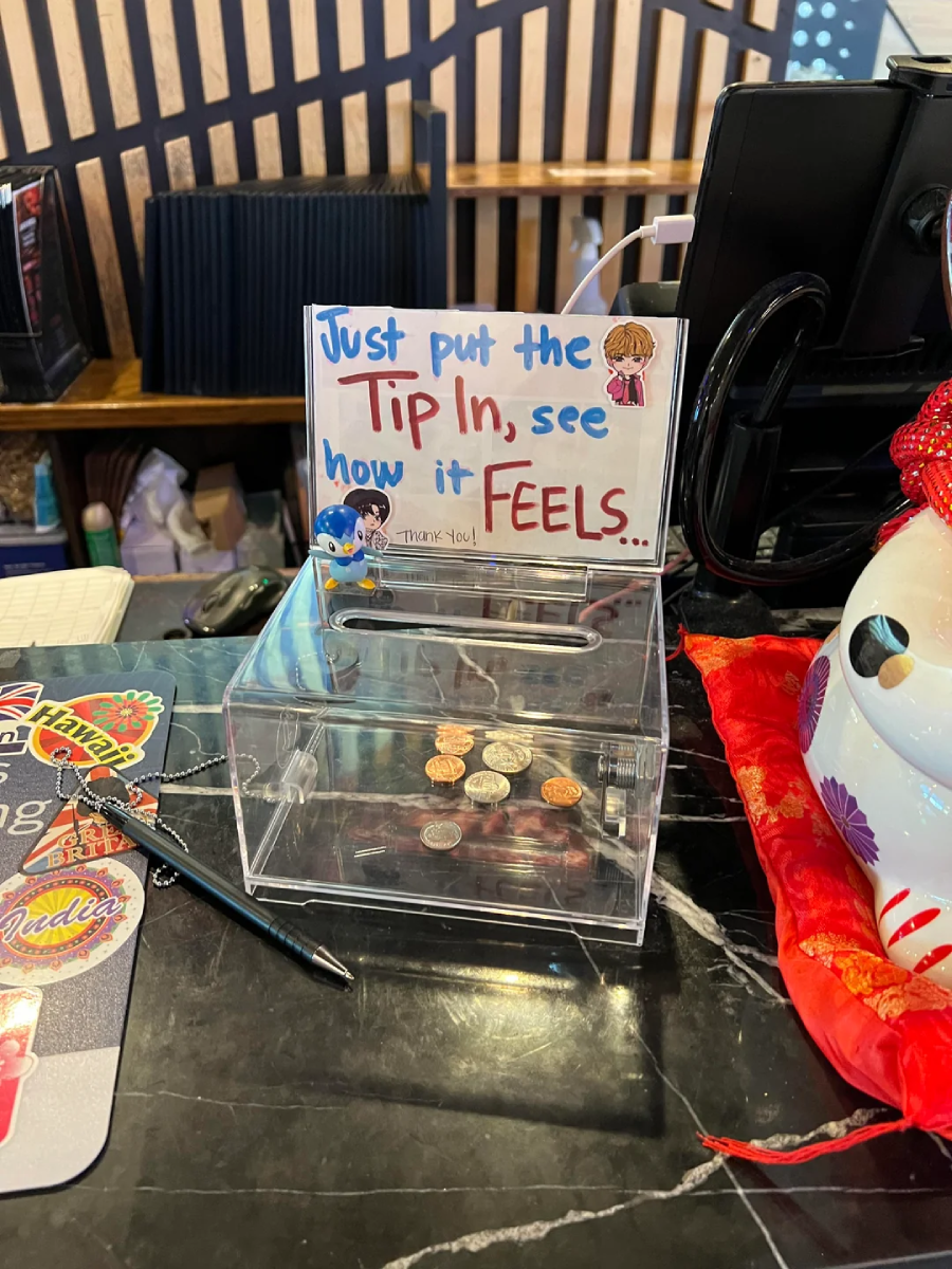 A clear tip box with coins inside sits on a counter. A colorful sign above reads, "Just put the tip in, see how it feels..." A pen and part of a decorated container are also on the black marble countertop.