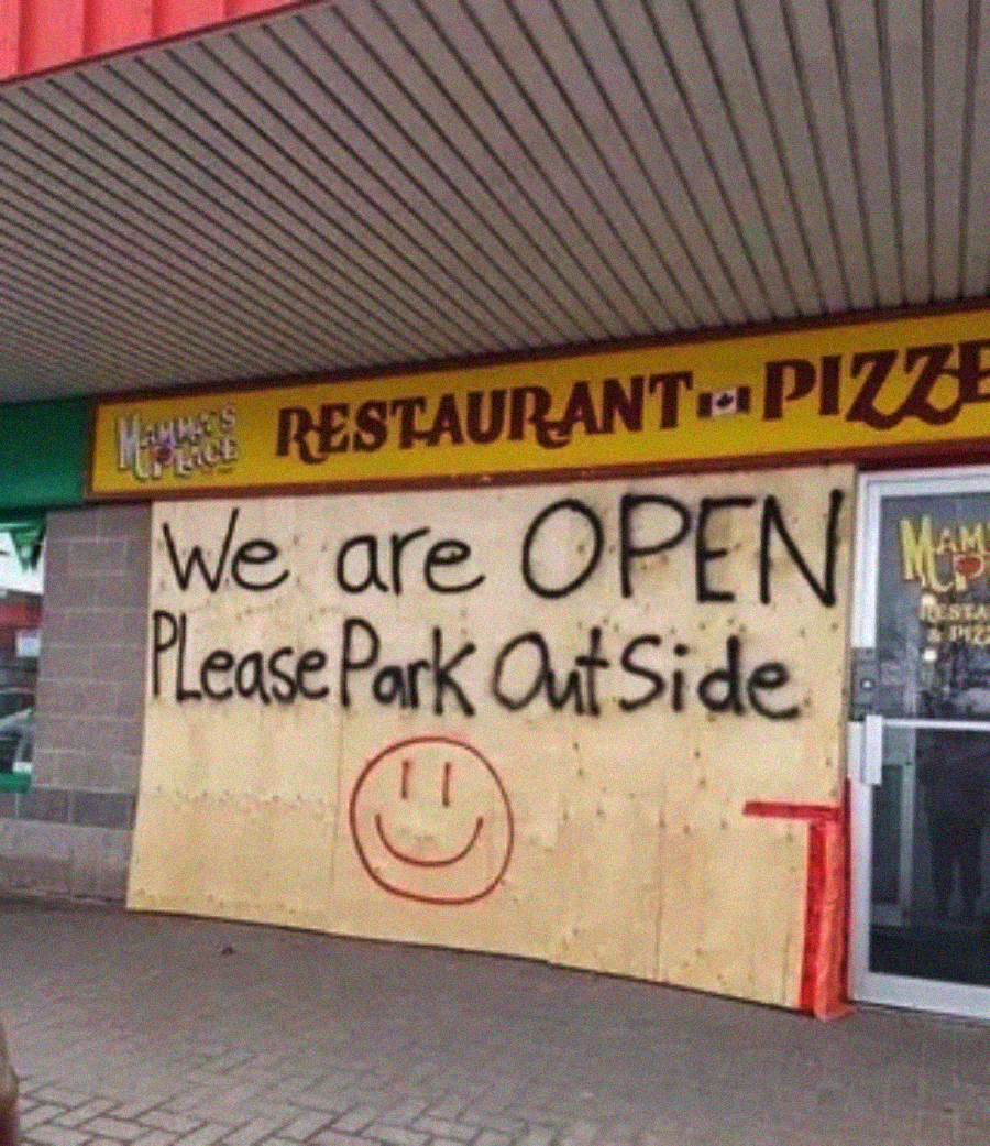 A restaurant with a large handwritten sign on plywood reads, "We are OPEN Please Park Outside" with a smiley face, placed over the front windows beside a glass door.