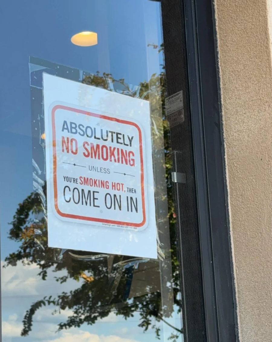 A sign on a glass door reads: “Absolutely no smoking unless you’re smoking hot, then come on in,” with a tree and sky reflected in the glass.