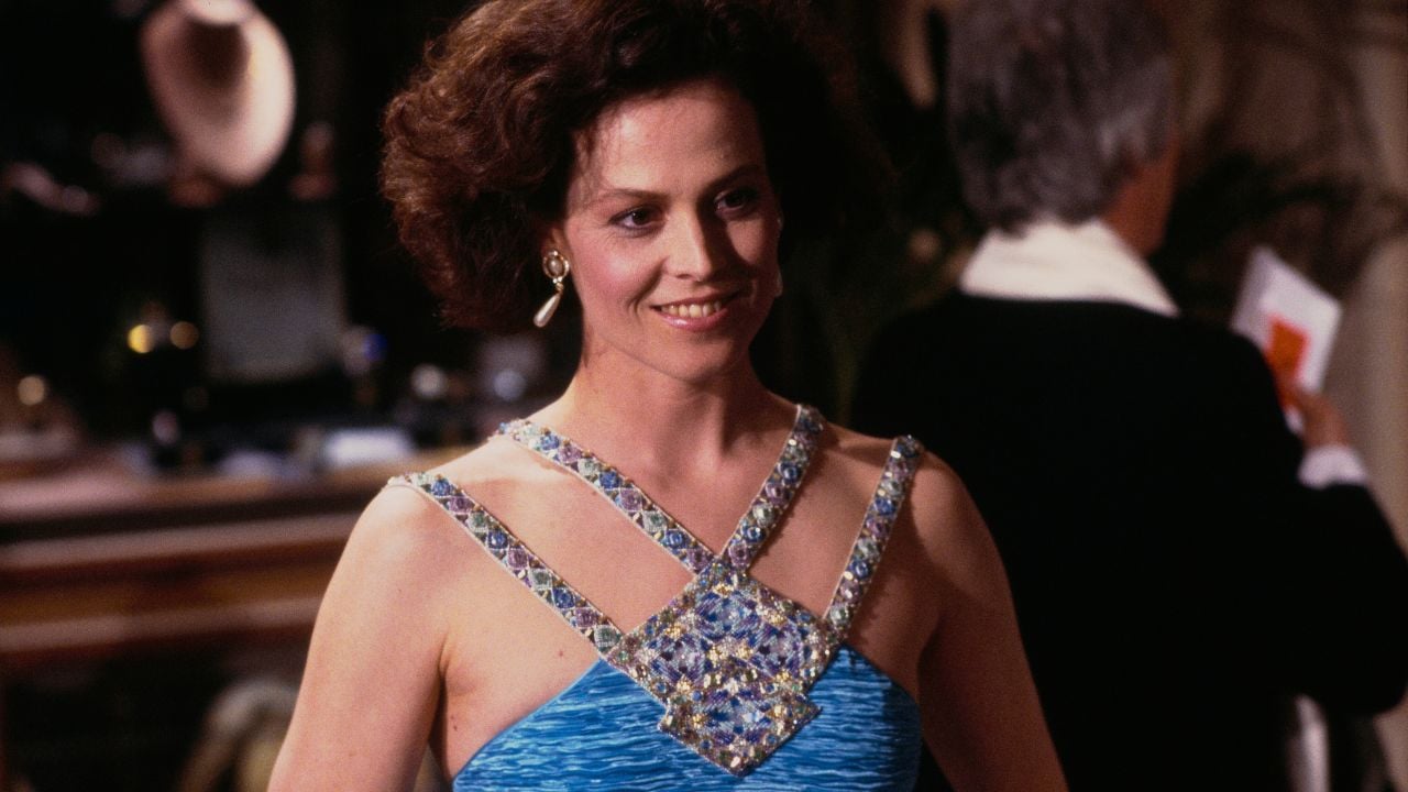 A woman with short, curly brown hair wears a blue dress with jeweled straps and smiles. She stands indoors, with a blurred background and another person visible behind her.