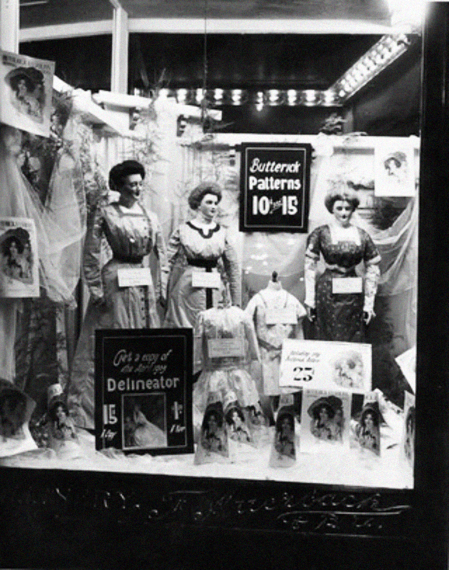 Black and white photo of a vintage store window display with mannequins in early 1900s dresses, signs for Butterick Patterns, and illustrations of women in similar outfits. Promotional materials are arranged around the mannequins.