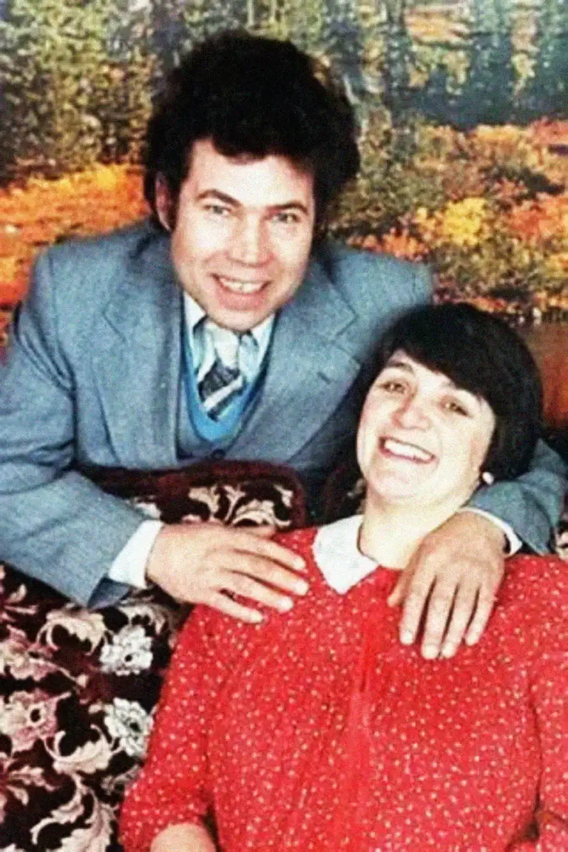 A man in a grey suit and blue tie stands behind a smiling woman in a red dress with white polka dots, resting his hand on her shoulder. They are posed in front of a scenic, autumn-themed backdrop.