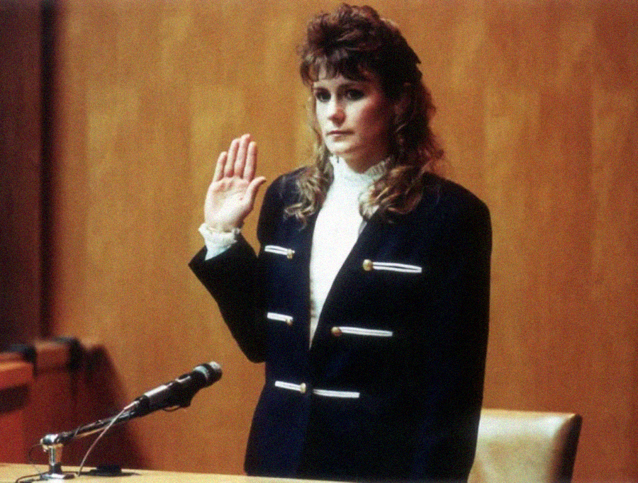 A woman in a black jacket with gold buttons stands in a courtroom, raising her right hand to take an oath while testifying. A microphone is visible on the desk in front of her.