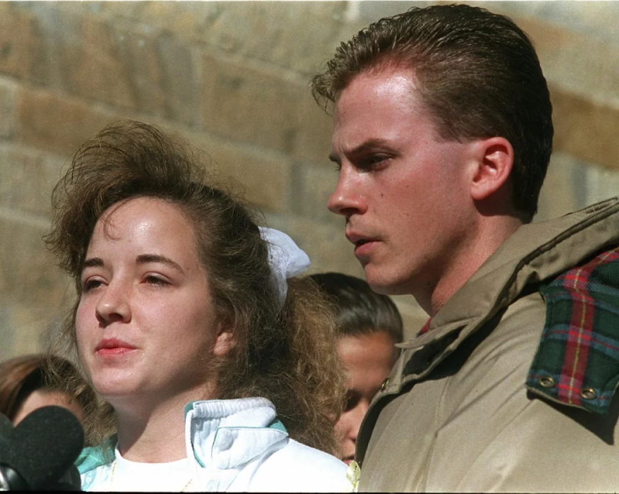 A woman and a man stand close together outdoors, both looking serious and focused. The woman wears a white jacket and has a white scrunchie in her hair. The man wears a beige coat. There are blurred people and a stone wall in the background.