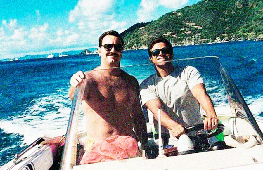 Two men smiling on a boat in sunny weather, with blue water and green hills in the background. One is shirtless with sunglasses and a mustache; the other wears a light t-shirt and sunglasses while steering the boat.