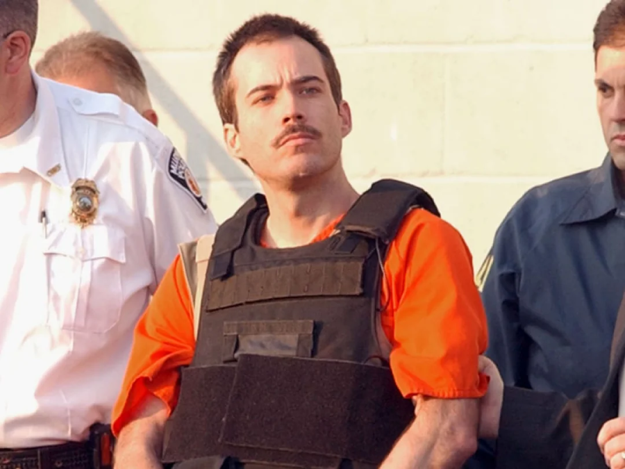 A man in an orange prison uniform and black protective vest is escorted by law enforcement officers, including one in a white shirt, standing against a light-colored wall.