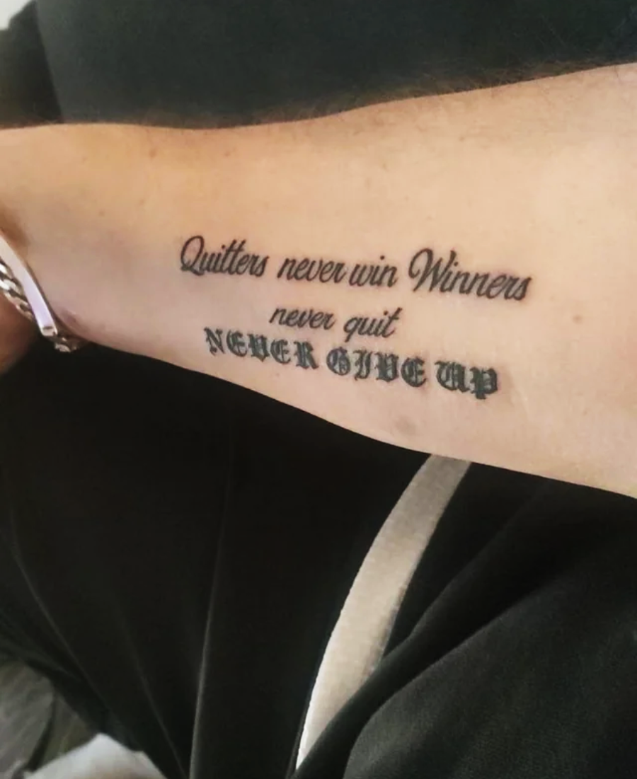 A tattoo on a person's inner arm reads: "Quitters never win Winners never quit NEVER GIVE UP" in different fonts and sizes.