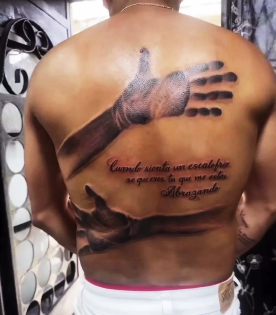 A person’s back displays a tattoo of two realistic arms and hands appearing to embrace them, with Spanish cursive text beneath the arms. The person is indoors and is shown from the waist up, facing away.