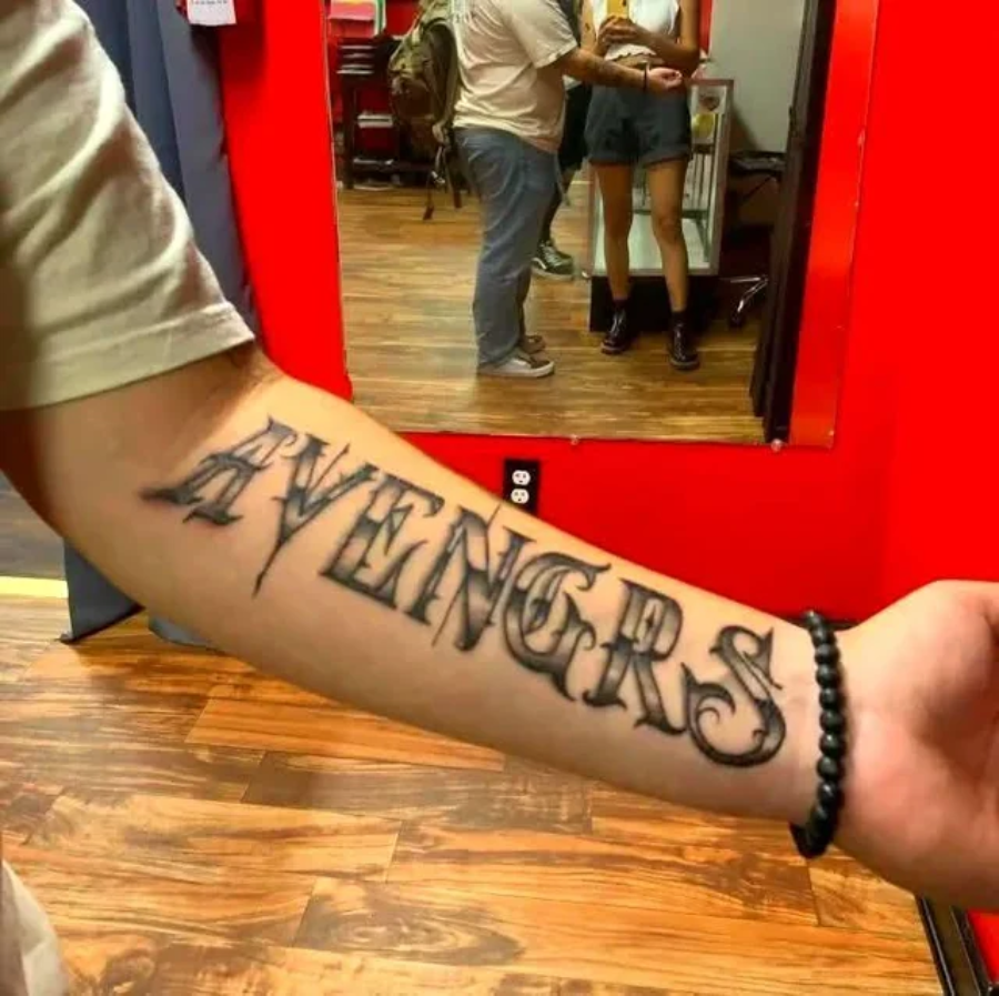 A person shows their inner forearm tattoo that reads "AVENGRS" in large, bold, stylized letters, missing the letter "E." The background features a red wall, a mirror, and a person with a camera.