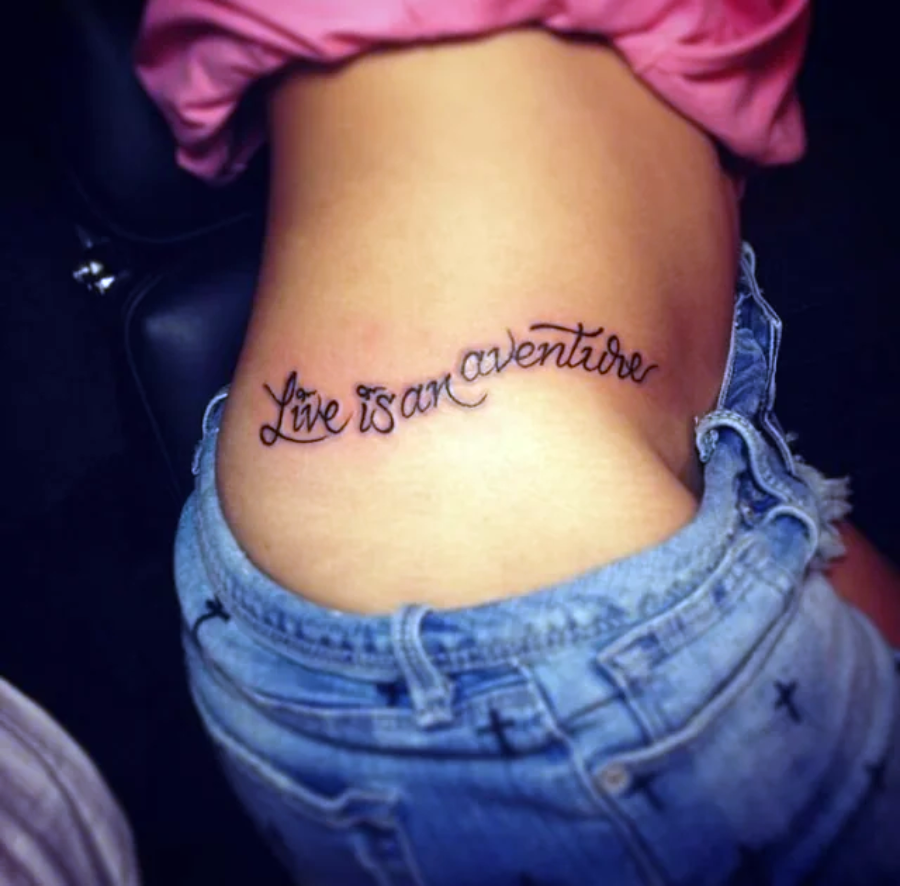 A person with denim shorts and a pink top shows a tattoo on their side reading "Live is an aventure" in cursive script.