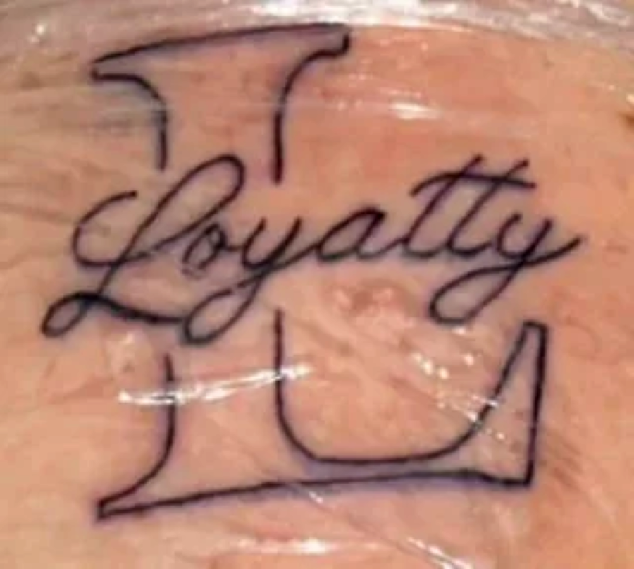 Tattoo of a large capital letter "L" with the word "Loyatty" written in cursive across it. The skin around the tattoo is covered in a clear protective wrap.