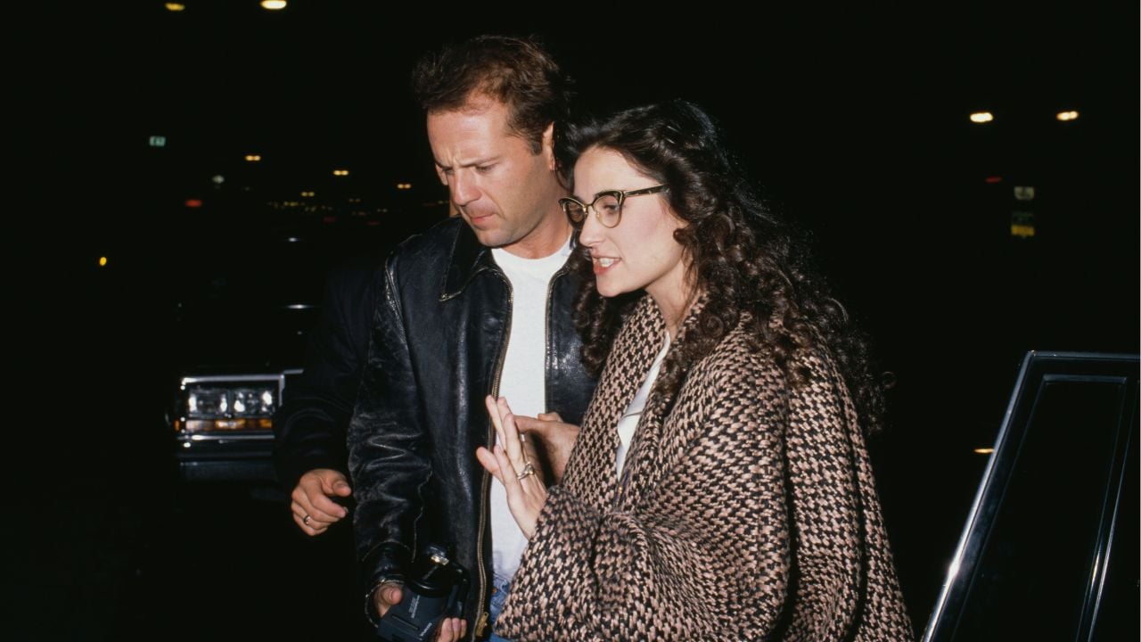 A man in a black leather jacket stands next to a woman in glasses and a brown patterned shawl at night, near a car with its door open. Both appear to be walking or leaving a vehicle.