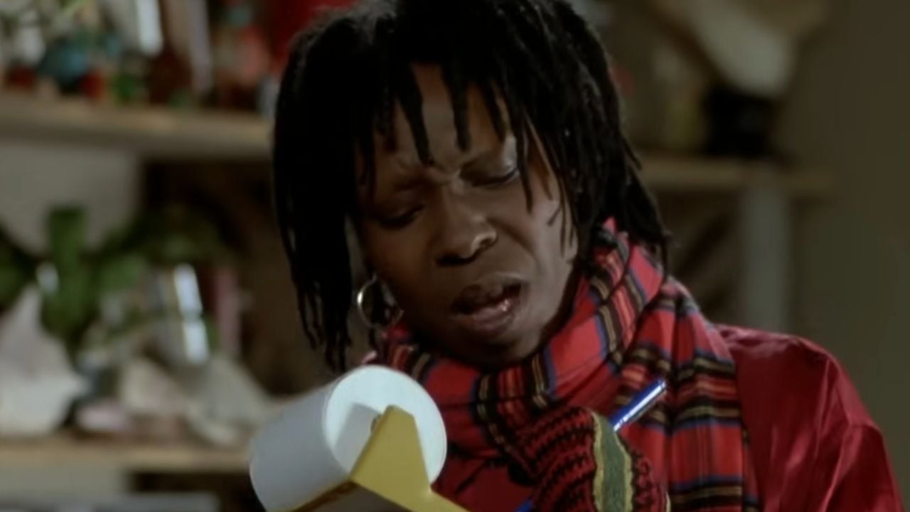 A person with dreadlocks, wearing a red and black checkered scarf and red clothing, looks at a notepad and pen, appearing focused and thoughtful in a cozy indoor setting.