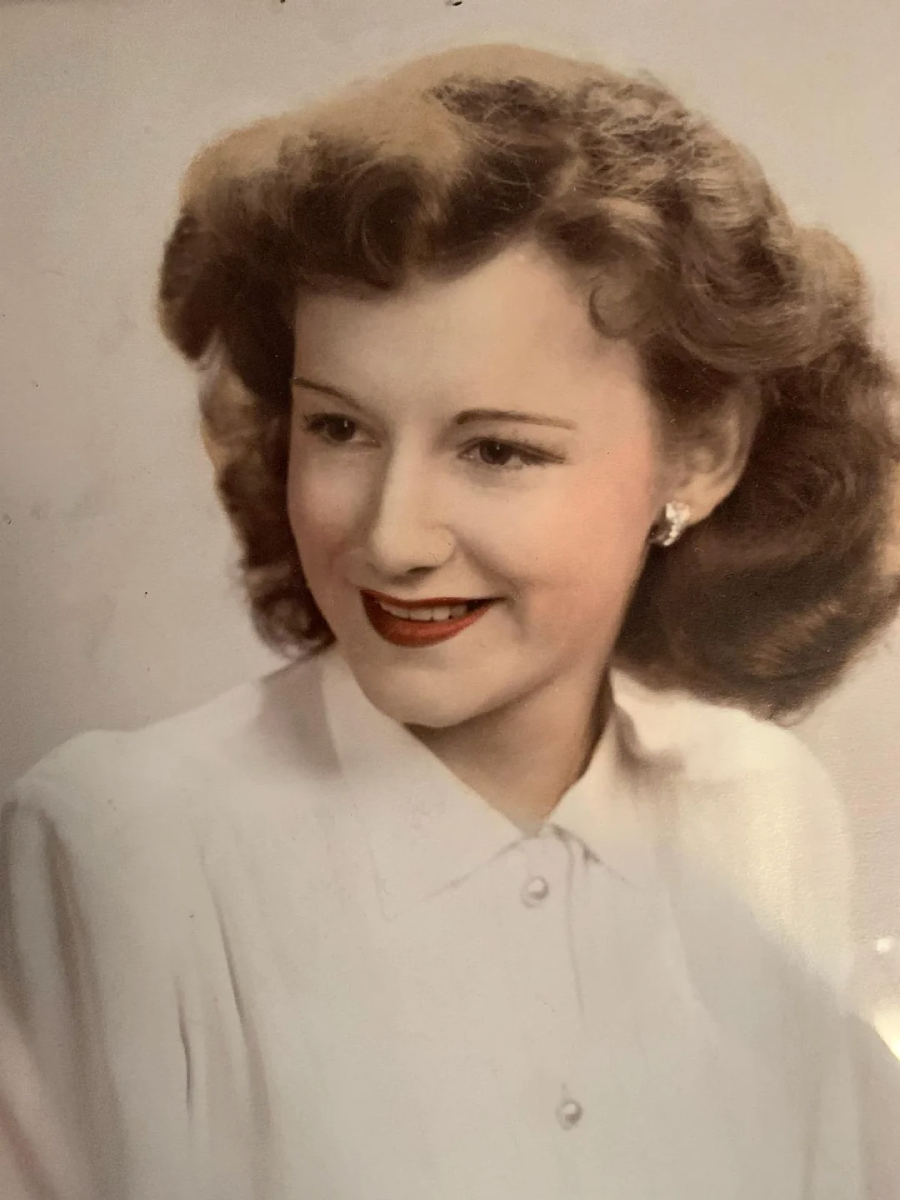 A young woman with wavy, light brown hair smiles slightly, wearing a white blouse with a buttoned collar and small earrings, against a plain background.