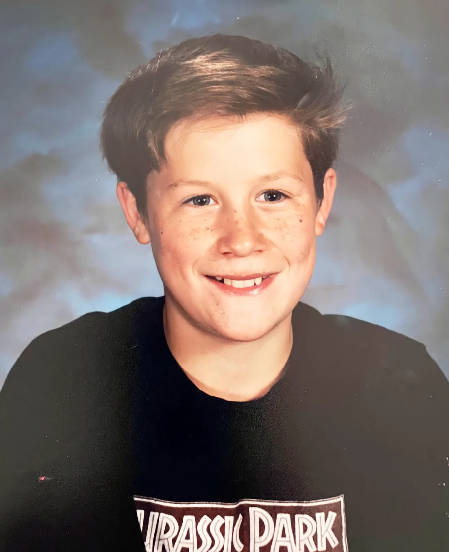 A smiling young boy with short brown hair and freckles wears a black Jurassic Park T-shirt in a classic school photo with a cloudy blue background.