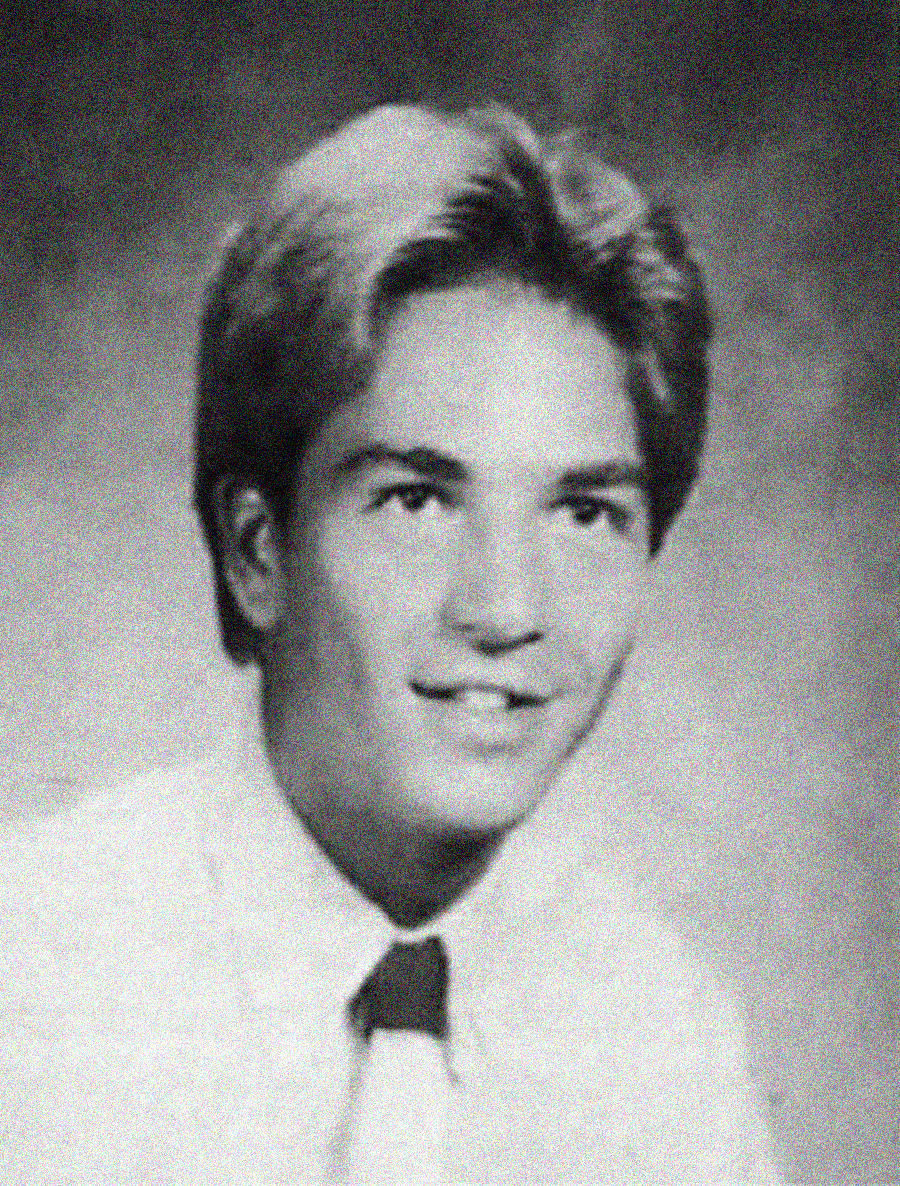 Black and white yearbook-style portrait of a young man with light hair, wearing a white shirt and a dark tie, smiling slightly and facing the camera against a plain backdrop.