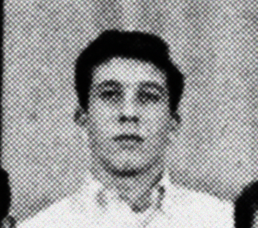 A grainy, black-and-white photo shows a young man looking directly at the camera. He has short dark hair and is wearing a collared shirt, with a neutral expression on his face against a plain background.