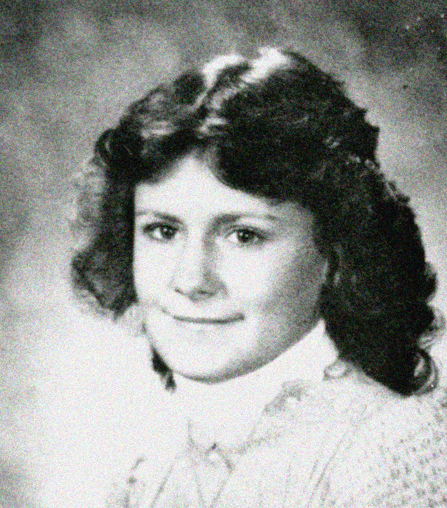 Black and white portrait of a young girl with wavy, shoulder-length hair, wearing a light-colored top with a high collar. She is facing the camera and smiling softly. The background is plain and slightly blurred.