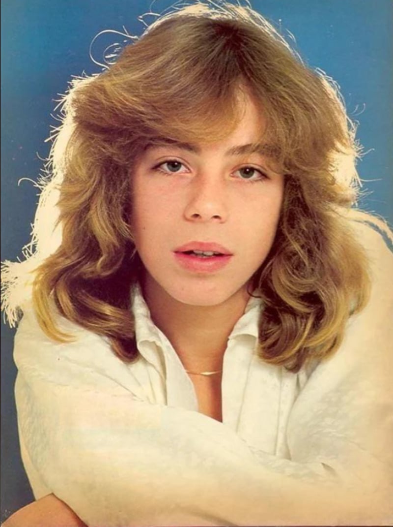 A young person with long, wavy blond hair and fair skin poses against a blue background, wearing a white shirt and looking directly at the camera with a neutral expression.