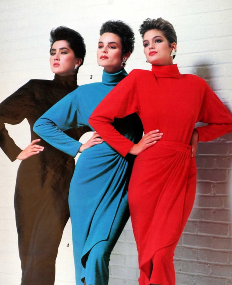 Three women pose in front of a white brick wall, wearing bold, long-sleeved dresses in black, teal, and red. The dresses have high necks and draped details, reflecting 1980s fashion styles.
