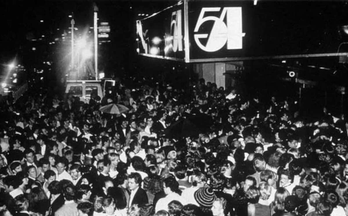 A large crowd of people gathers outside the entrance of Studio 54 at night, under a brightly lit sign displaying the club's name and number. The scene is lively and packed, capturing the excitement of the nightclub.