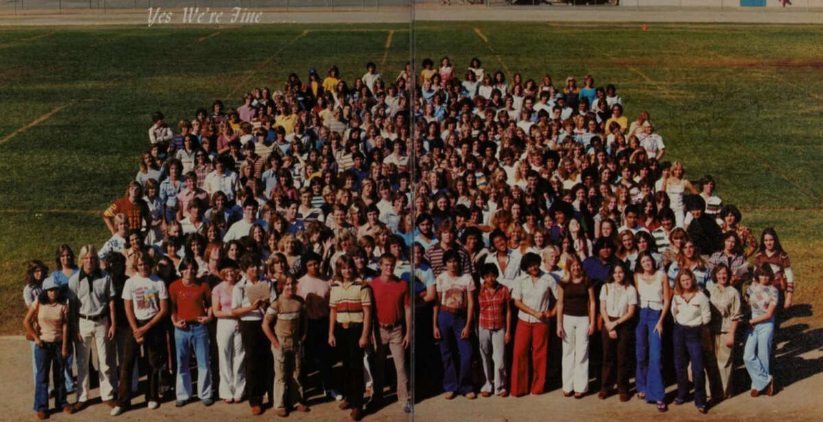 A large group of high school students stands closely together outdoors on a grassy field, posing for a group photo. Text at the top reads, “Yes We’re Fine.”