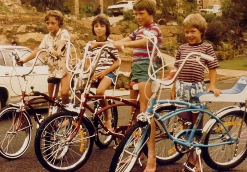 Four children stand outside with vintage bicycles, smiling and dressed in striped shirts and shorts. A white car and rocky landscape can be seen in the background. The scene has a retro, nostalgic feel.