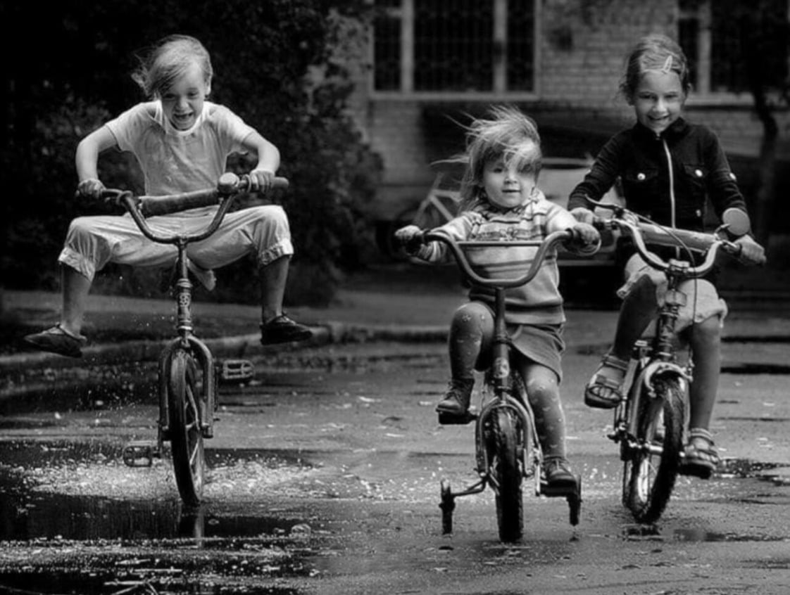 Three young children ride bicycles through a puddle on a street. Two laugh joyfully while the third looks ahead, all with windblown hair and wet clothes, capturing a moment of carefree play. The image is in black and white.