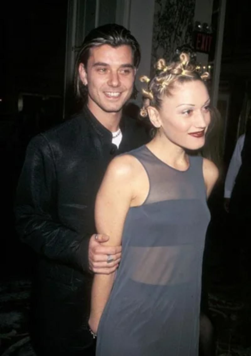 A smiling man in a dark jacket stands behind a woman with blonde Bantu knots and dark lipstick, wearing a sheer gray sleeveless dress, at an indoor event.
