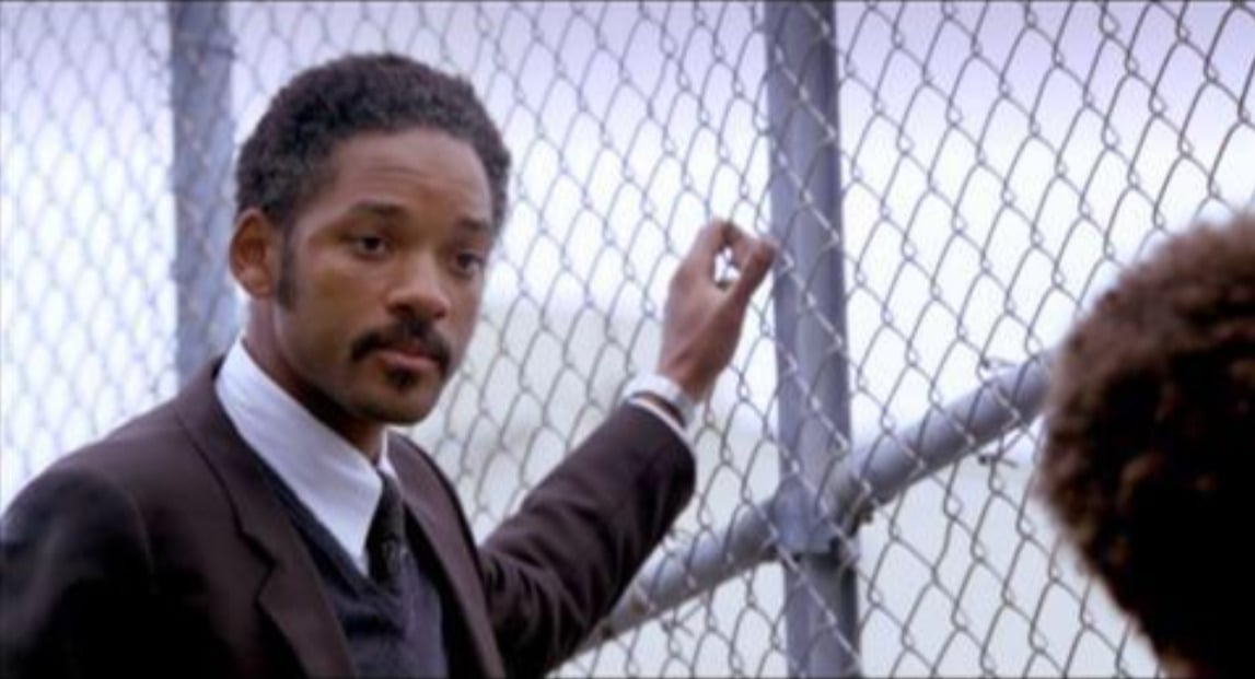 A man in a suit stands beside a chain-link fence, looking thoughtfully at someone out of frame. The background is blurred, and the sky appears overcast.