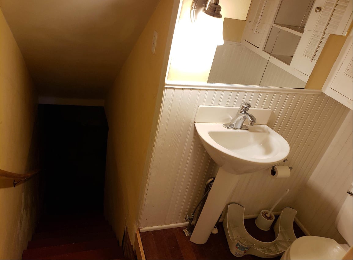 A small bathroom with a pedestal sink and mirror is situated at the top of a dark, narrow staircase, with a light illuminating the sink area and a toilet seat on the floor nearby.