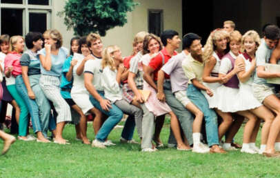 A group of smiling teenagers stands outside on grass, lined up and leaning on each other in a playful, zigzag pose. They wear casual summer clothes and appear to be enjoying a group activity.