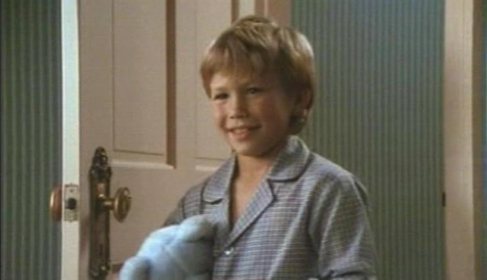 A young boy with blond hair, wearing blue checkered pajamas, stands and smiles while holding a blue stuffed animal, with a white door and green striped wall in the background.