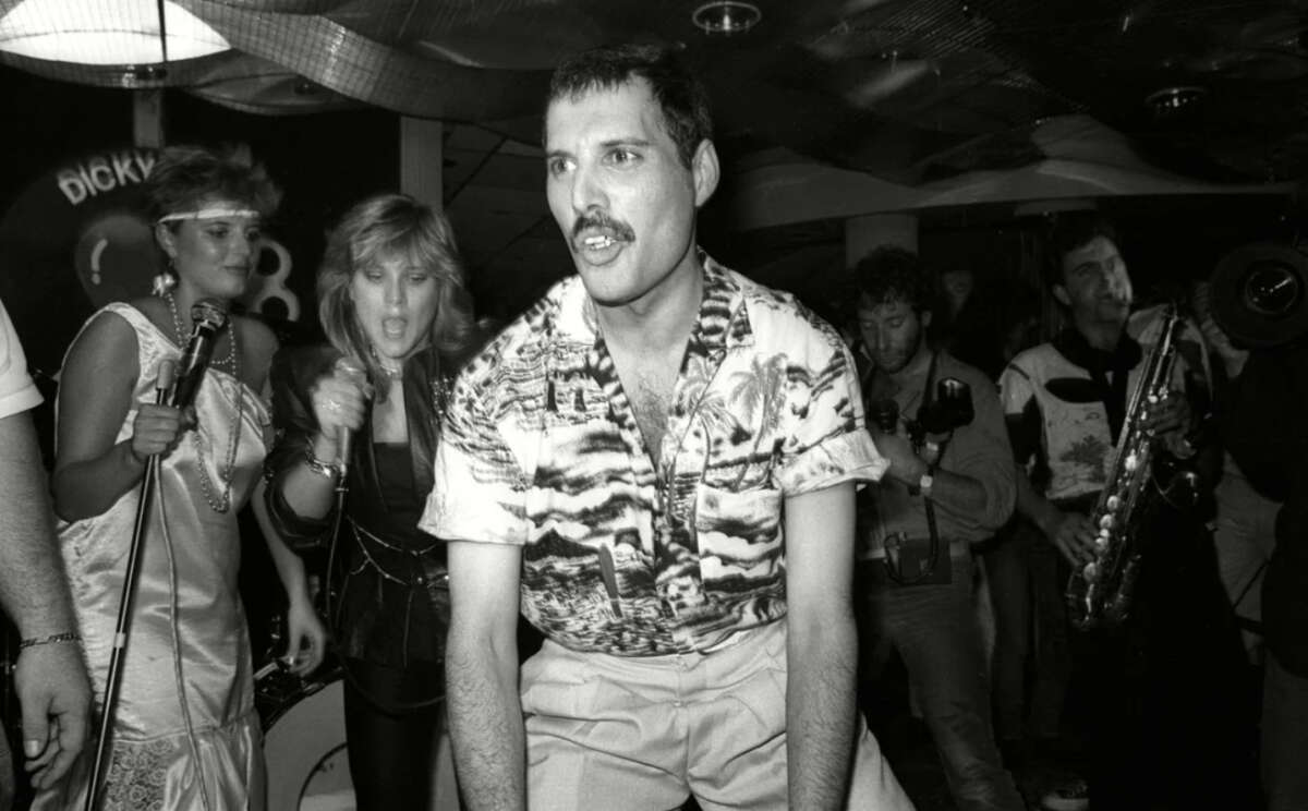 A man in a patterned shirt dances energetically at a lively party, surrounded by people singing, playing music, and enjoying the festive atmosphere. The setting appears to be indoors with musical instruments visible.