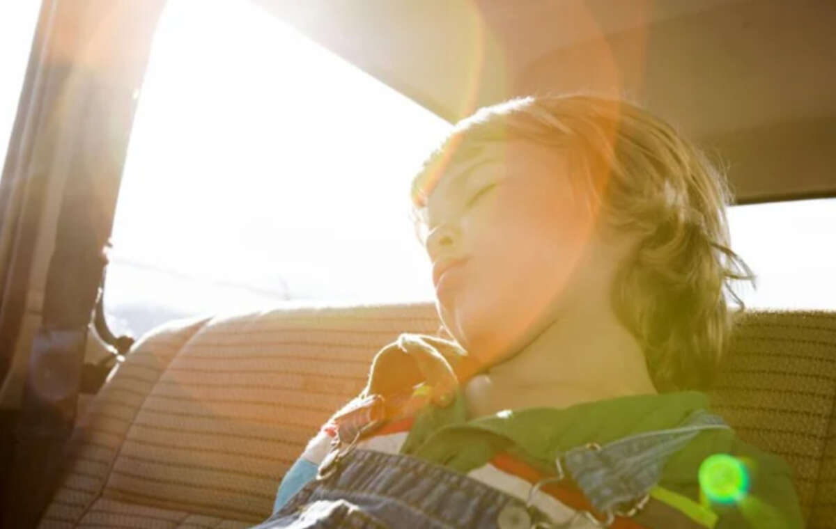 A young child with light hair is sleeping in the back seat of a car, bathed in warm sunlight with lens flare visible. The child is leaning back, wearing a green shirt and blue overalls, and appears peaceful.