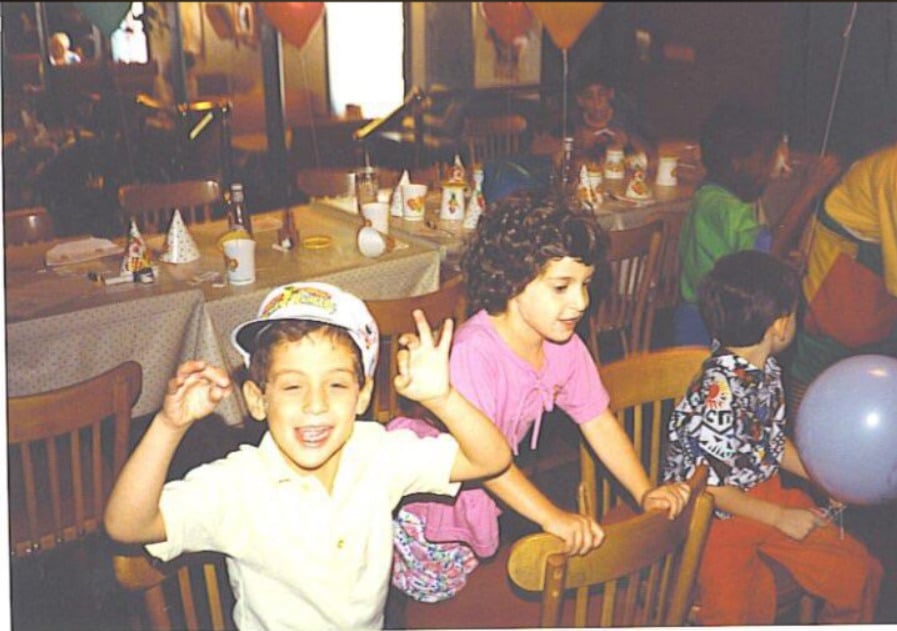 Children smile and play around a table set for a party with hats, cups, and balloons. One boy in front waves with both hands, and another child wearing pink stands nearby. The room is decorated for a celebration.