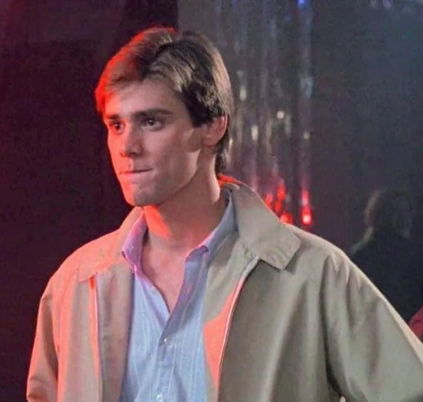 A young man with light brown hair wearing a beige jacket over a blue button-down shirt stands indoors with a serious expression, lit by red and blue lighting in the background.