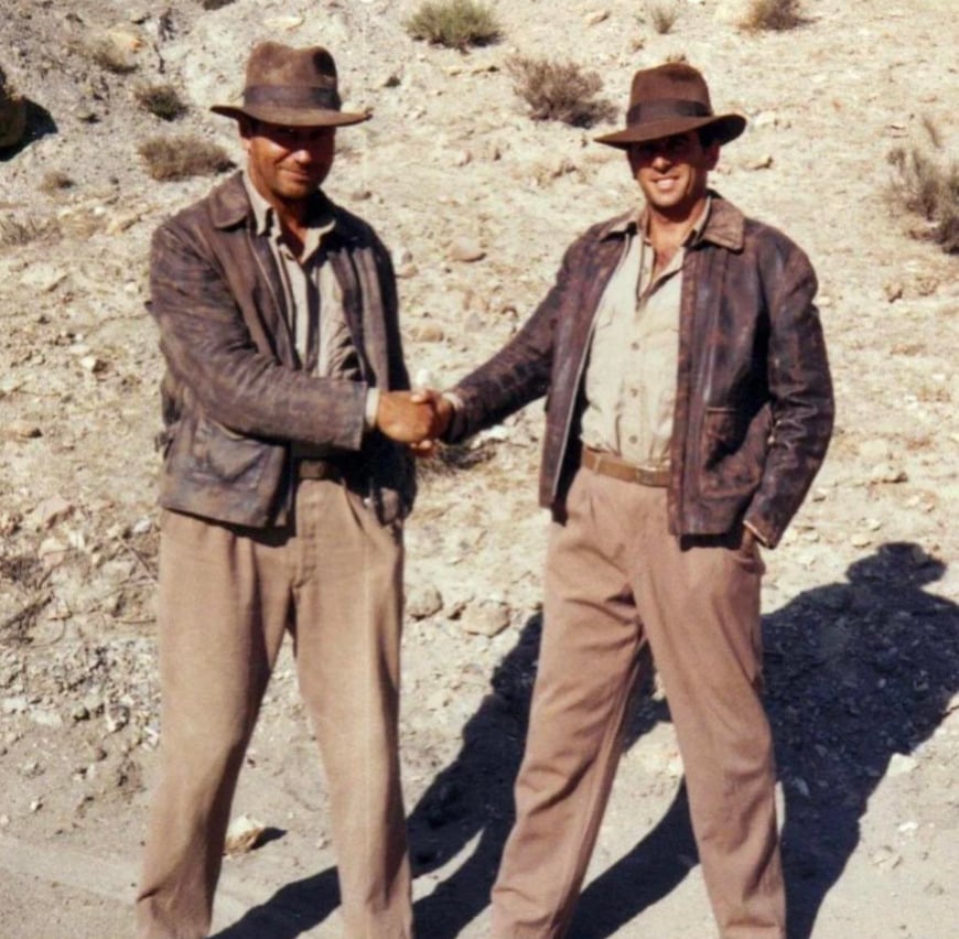 Two men wearing brown fedoras, leather jackets, and beige pants shake hands outdoors on a dry, rocky terrain. Both are smiling, and the sunlight casts their shadows on the ground.