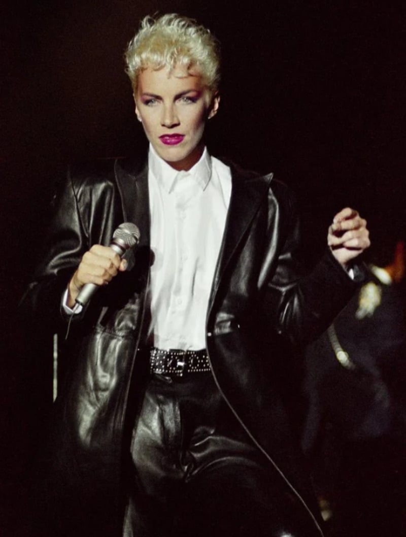 A person with short, platinum blonde hair wears a black leather suit and white shirt, holding a microphone and performing on stage under dramatic lighting.