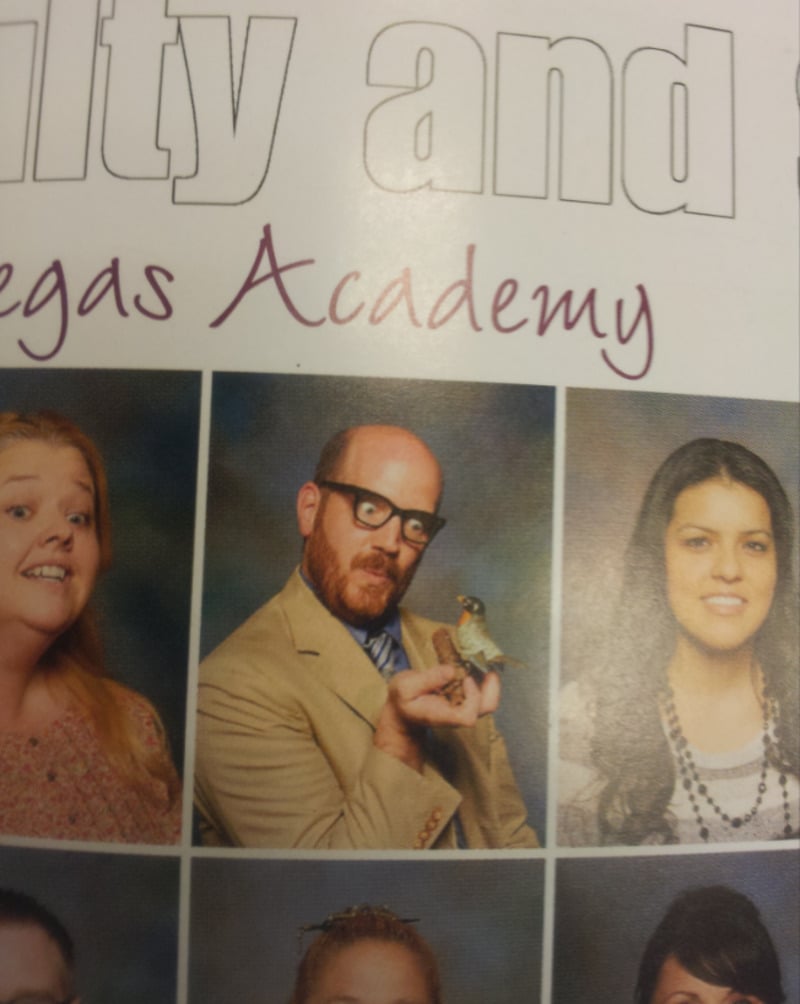 A man wearing glasses and a tan suit poses for a school photo while holding a small bird; he is flanked by two women, all in front of a multicolored backdrop with text partially visible above.