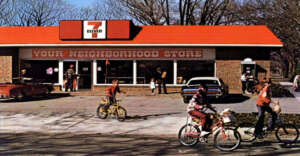 Children ride bicycles in front of a vintage 7-Eleven store with a red roof and a sign that reads "YOUR NEIGHBORHOOD STORE." Two classic cars are parked, and trees without leaves are in the background.