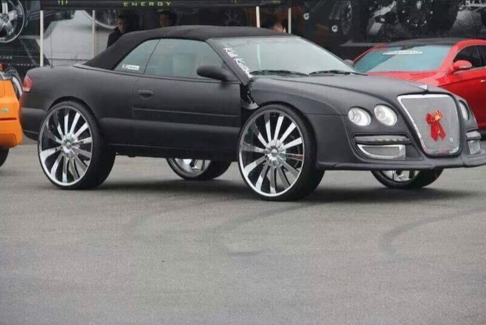 A matte black convertible car with extremely large, shiny chrome rims and low-profile tires is parked at a car show. The car has an unusual, custom luxury front grille with a red bow on it.