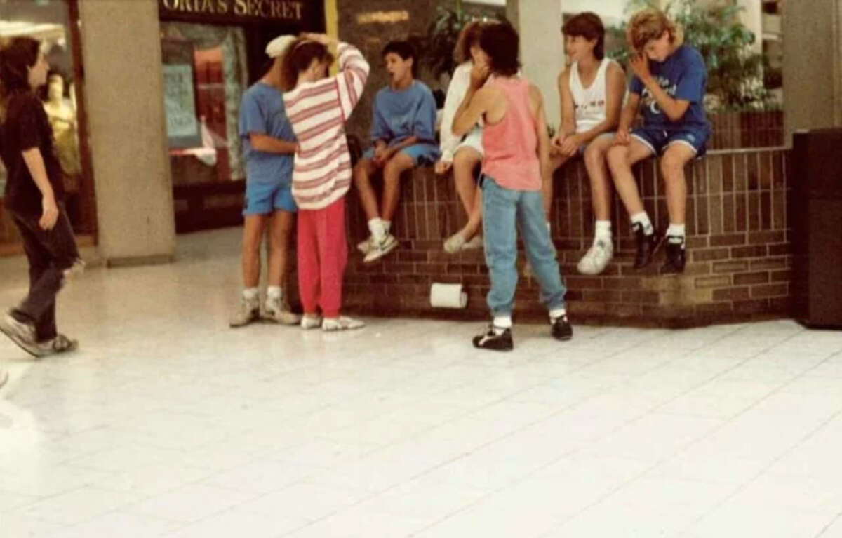 A group of kids in casual 80s-style clothes gather and sit on a low brick wall in a mall, talking. A store with a “Victoria’s Secret” sign is visible in the background. The floor is tiled white.