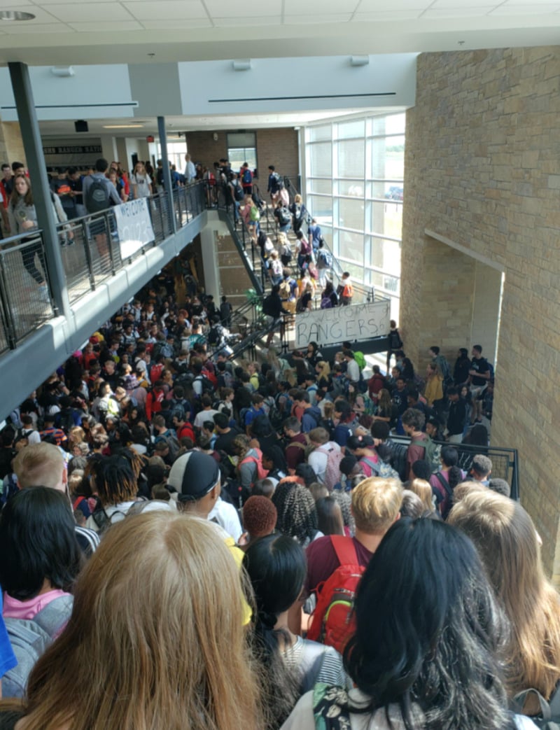 A large crowd of students with backpacks fills a busy school hallway and staircase, gathering below a banner that reads “RANGERS.” Sunlight streams in through tall windows on the right side.