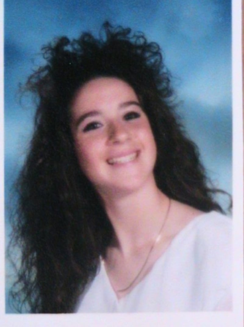 A young woman with long, curly dark hair and a big smile poses for a portrait against a blue background. She is wearing a white top and a delicate gold necklace.