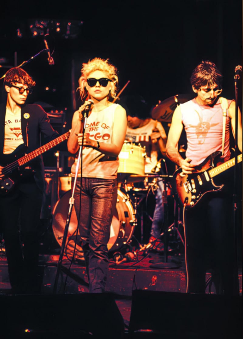 A band performs on stage; the singer in the center wears sunglasses and a graphic tank top, flanked by two guitarists, with a drummer visible in the background under colorful stage lighting.
