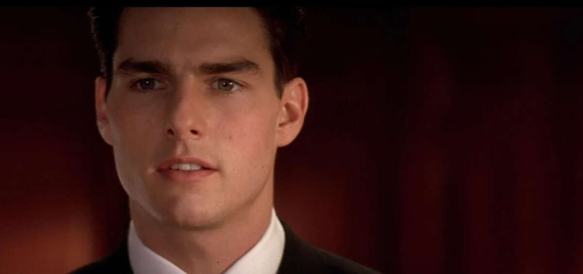 A man in a suit and tie looks ahead with a serious expression against a dark, blurred background.